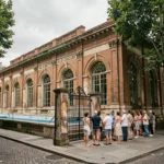 File d'attente devant la piscine de la Butte aux Cailles à Paris, façade Art Déco en briques avec grille en fer forgé et visiteurs en tenue estivale