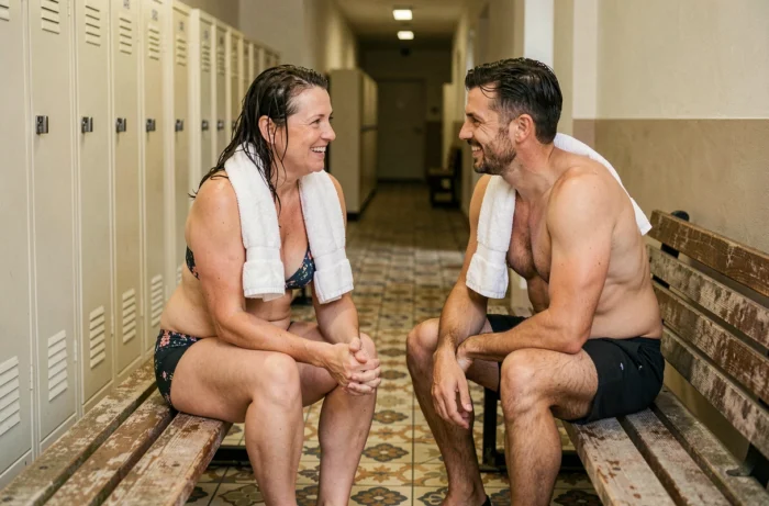 Deux nageurs souriants assis sur des bancs en bois dans le couloir des vestiaires de la piscine de la Butte aux Cailles après leur séance de natation